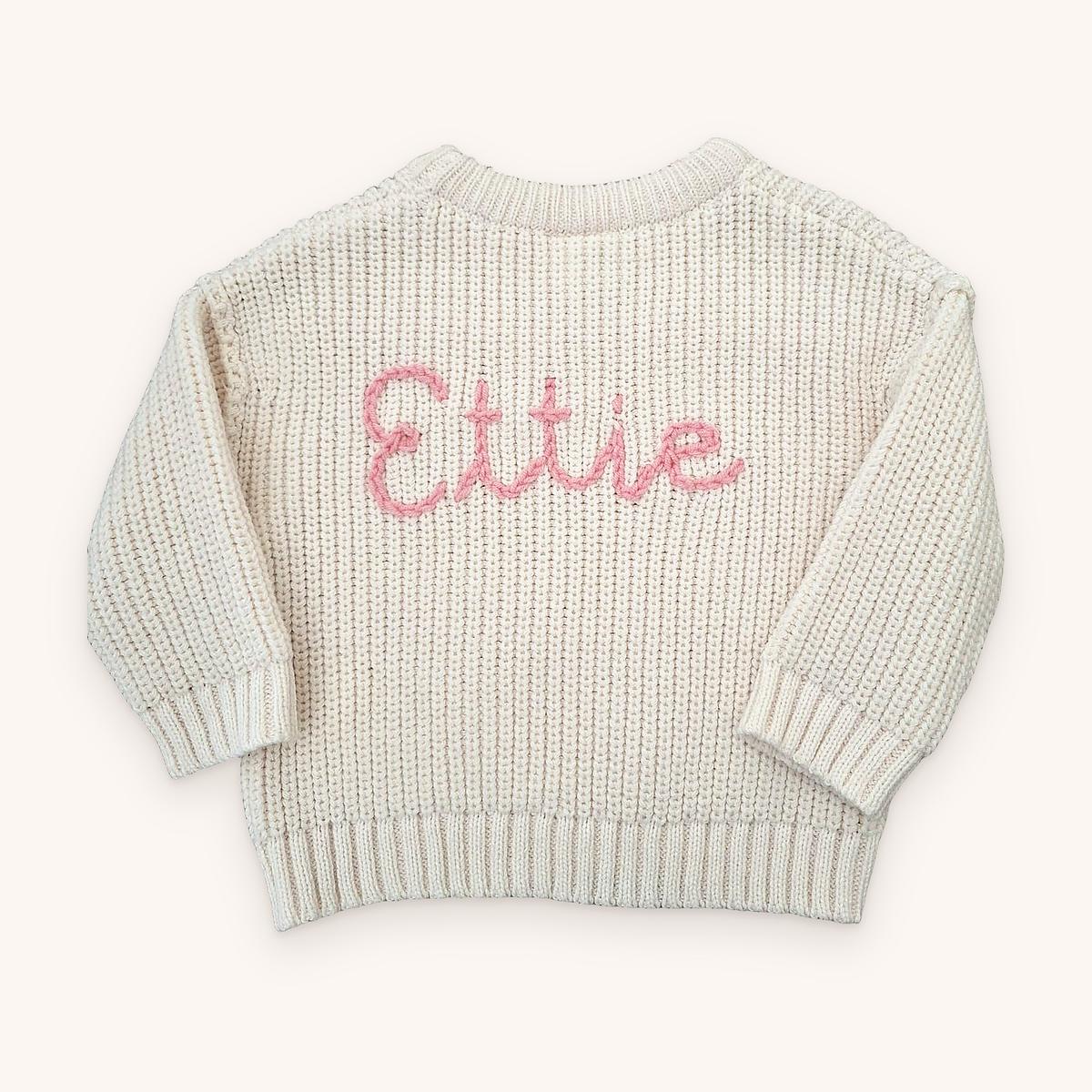 Cream chunky knit baby jumper with the name Ettie hand-embroidered across the front in pink chain-stitch script
