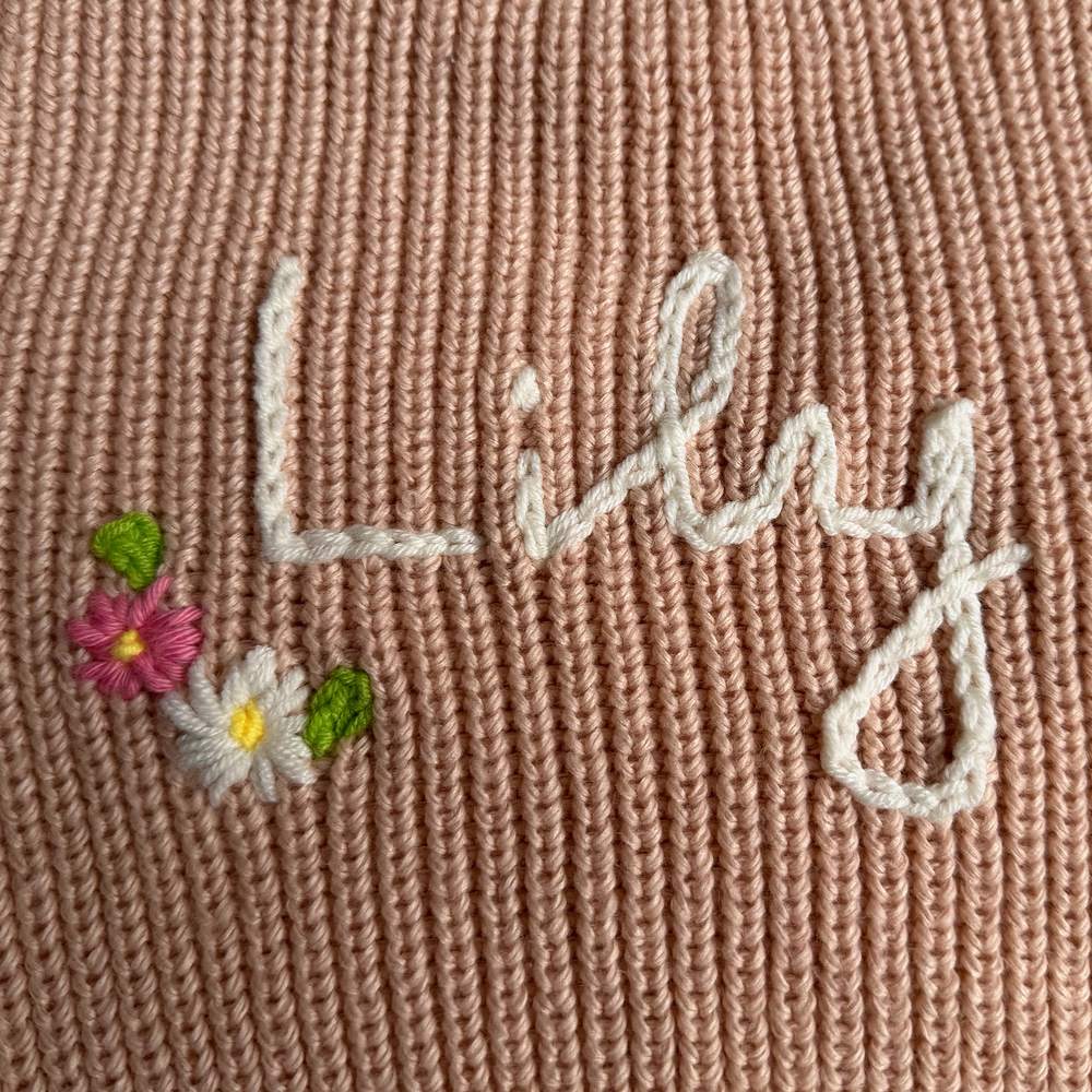 Close-up of the hand-stitched cream chain-stitch name Lily on a dusty pink chunky knit, with embroidered pink and white flowers and green leaves